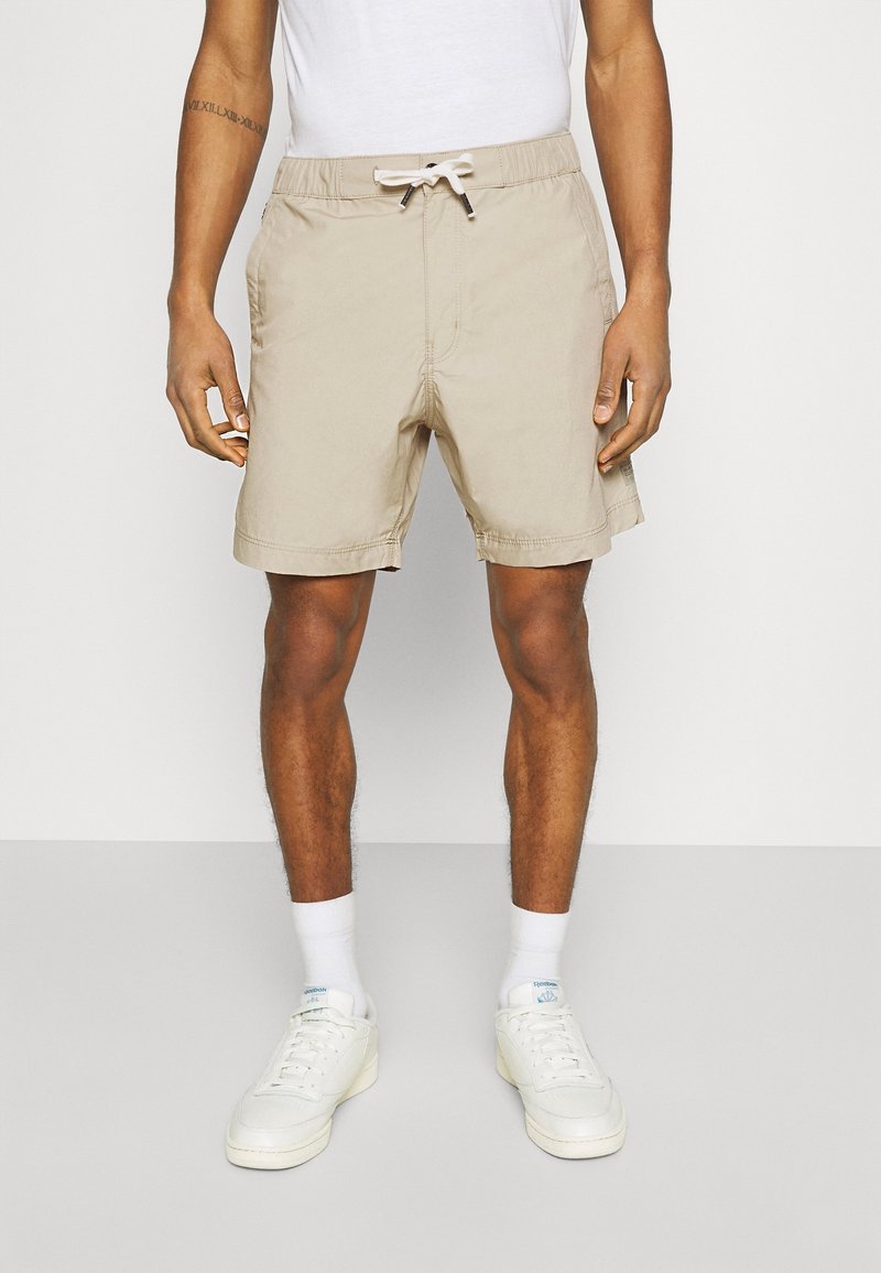 Man wearing beige drawstring shorts, white socks, and white sneakers standing against a plain background.