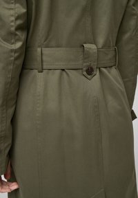 Cappotto verde oliva in tessuto testurizzato, con vita accentuata da una cintura, dettagli con bottoni e cuciture a vista sul retro.