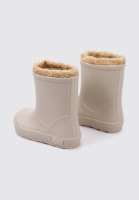 Botas de goma beige con un suave forro de pelaje sintético en tono marrón, punta redonda, textura acanalada y suela texturizada. Logo estampado en el talón.