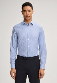 Camicia button-up azzurro chiaro con un piccolo motivo a quadri, maniche lunghe e colletto aperto. Indossata infilata in pantaloni scuri.