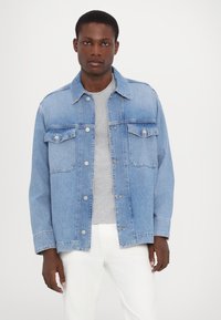 Light blue denim jacket with a classic collar, two chest pockets, and silver snap buttons. Worn over a grey shirt and white pants.
