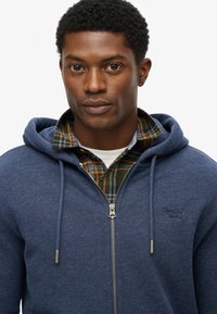Giovane uomo che indossa una felpa blu con zip sopra una camicia a quadri verde e arancione, che guarda dritto davanti a sé con un'espressione neutra su uno sfondo bianco.