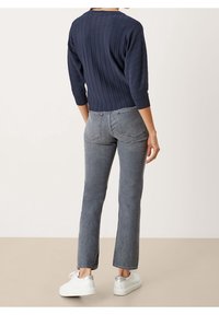 Personne portant un pull côtelé marine, un jean gris à jambes droites et des baskets blanches, debout contre un fond clair, vue de dos.