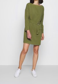 Olivgrön klänning med lös passform, långa ärmar, ett midjebälte med en silverring och en framficka; bärs med vita sneakers.