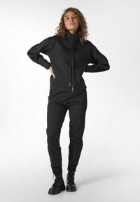 Zwarte sweatshirt met hoge kraag en rits, gecombineerd met aansluitende zwarte en groene streepbroek. Model draagt zwarte veterschoenen.