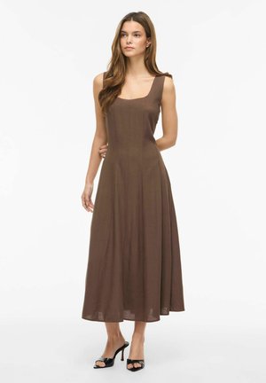 Femme aux longs cheveux bruns portant une robe midi marron sans manches et des sandales à talons noires, debout devant un fond blanc uni.