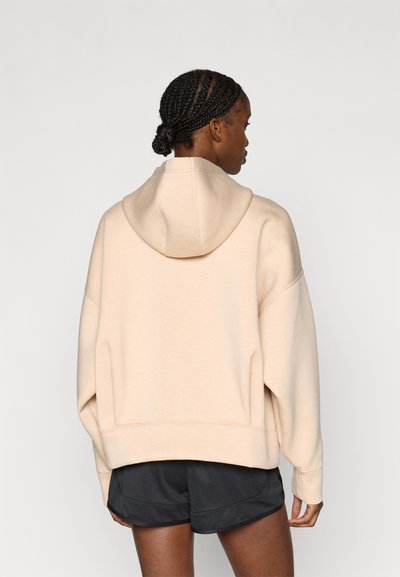 Μπεζ φαρδιά φούτερ με κουκούλα, με άνετη εφαρμογή και χαμηλωμένους ώμους. Συνδυάζεται με σκούρα σορτς, απεικονίζεται από πίσω.