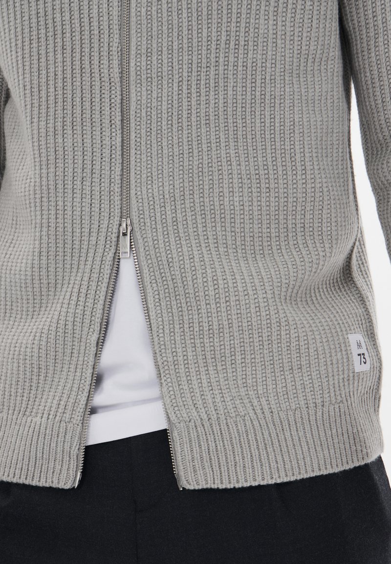 Grau gerippter Cardigan mit Reißverschluss vorne, seitlichen Schlitzen am Saum und einem kleinen Textillabel mit der Nummer 73. Leichte Stricktextur.