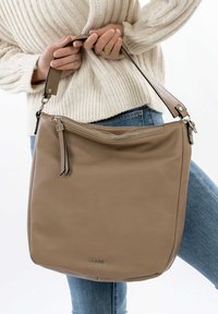 Sac à main en cuir beige avec une texture lisse, doté d'une fermeture éclair supérieure, d'une bandoulière réglable et d'un logo subtil à l'avant.