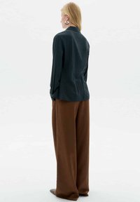 Frau mit blonden Haaren, die große runde Ohrringe, ein dunkles Nadelstreifen-Blazer und weite braune Hosen trägt und mit dem Rücken zu einem weißen Hintergrund steht.