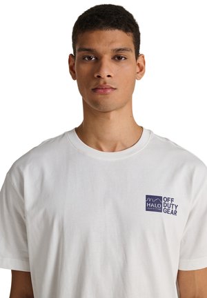 Jonge man met kort krullend haar, draagt een wit T-shirt met ronde hals en het logo "HALO OFF DUTY GEAR" op de linkerborst, neutrale uitdrukking, witte achtergrond.