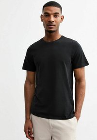 Camiseta de algodón negra, de manga corta, cuello redondo, textura suave, diseño ajustado, usada con pantalones cortos de color claro, fondo liso.