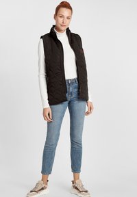 Svart quiltad väst med hög krage, dragkedja och sidofickor, bärs över en vit polotröja och blå skinny jeans.