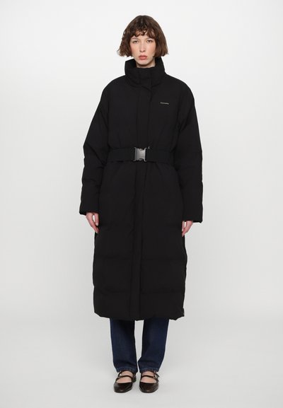 Long manteau rembourré noir à col haut, taille ceinturée et design matelassé. Associé à un jean bleu et des chaussures marron. Marquage minimaliste visible.