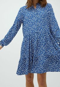 Blaues Kleid mit weißem Blattmuster, lange Ärmel, Knopfverschluss vorne, gestuftem Saum, aus einem leichten, strukturierten Stoff gefertigt.