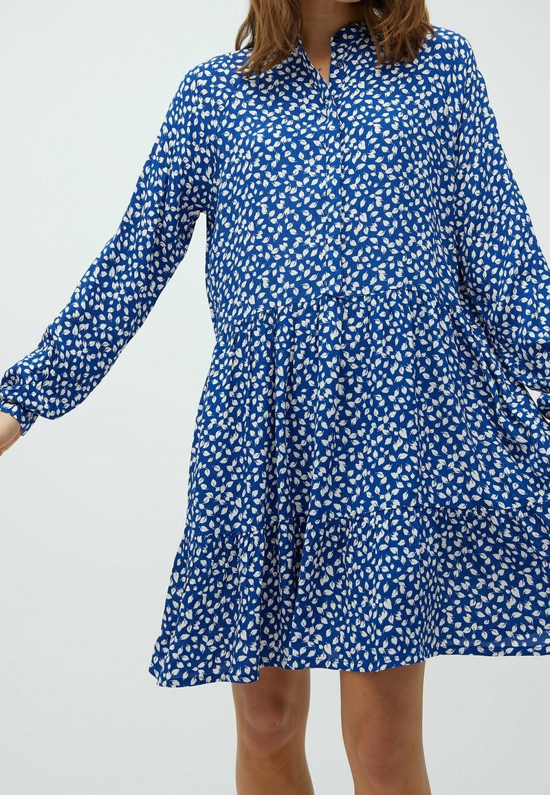 Blaues Kleid mit weißem Blattmuster, lange Ärmel, Knopfverschluss vorne, gestuftem Saum, aus einem leichten, strukturierten Stoff gefertigt.