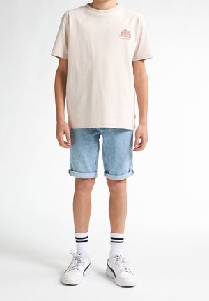 Jeune personne portant un t-shirt beige clair avec un petit logo, un short en denim bleu clair retroussé, des chaussettes blanches avec des rayures noires et des baskets blanches.