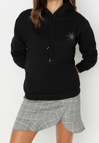 Sudadera negra con capucha, que presenta un gráfico decorativo de cuentas y un acento rojo. Combinada con una mini falda de cuadros en blanco y negro.