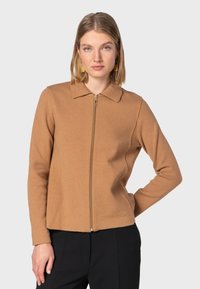 Beige zip-up jakke med ribbet krave og struktureret stof. Har lange ærmer og en tætsiddende pasform, parret med sorte bukser.
