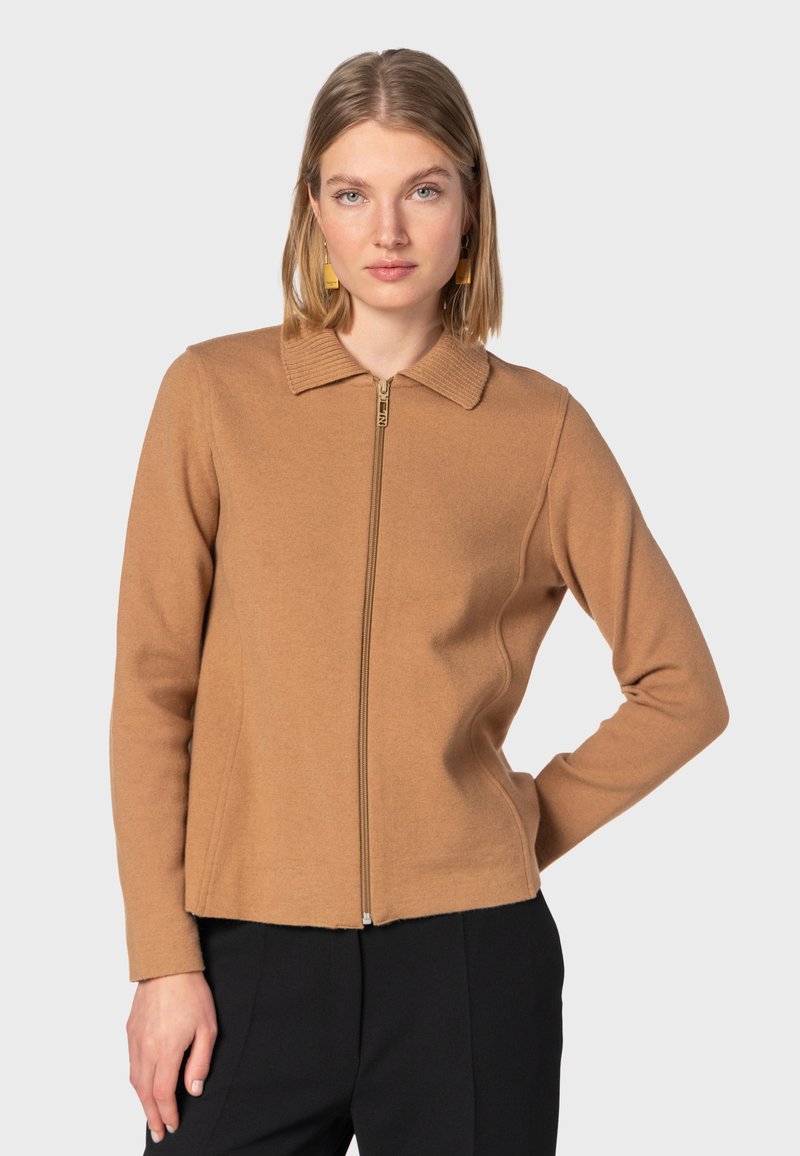 Beige zip-up jakke med ribbet krave og struktureret stof. Har lange ærmer og en tætsiddende pasform, parret med sorte bukser.