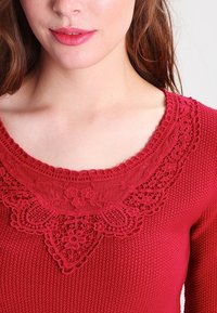 Pull en tricot rouge avec un col en dentelle détaillé et des motifs floraux, texture côtelée et une coupe légèrement cintrée.