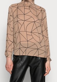 Blouse à manches longues en beige clair avec un motif géométrique noir, dotée d'un col montant froncé et d'un tissu doux et léger.