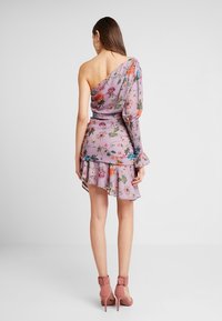 Žena v lila květinových šatech na jedno rameno s volánkovým lemem a nařasenými dlouhými rukávy, v kombinaci s růžovými sandály na vysokém podpatku, stojí s pohledem zezadu.