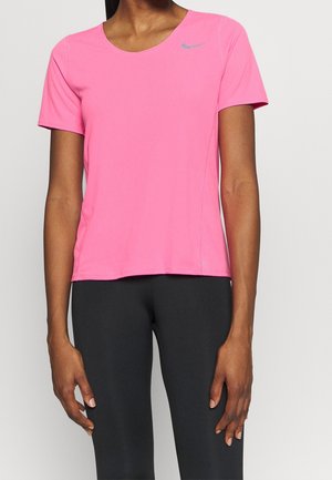 Femme portant un t-shirt de sport Nike manches courtes rose vif et des leggings noirs, debout devant un fond clair uni.