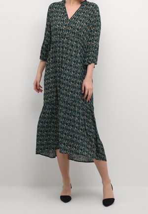 Femme portant une robe mi-longue vert foncé à motif floral et manches trois-quarts, associée à des talons noirs à bouts pointus.