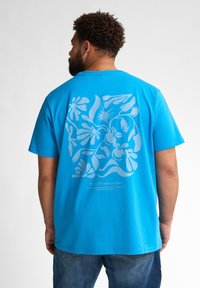 Modrá bavlněná tričko s velkým světle modrým grafickým designem na zadní straně, obsahující abstraktní tvary a rostliny. Volný střih, krátké rukávy.