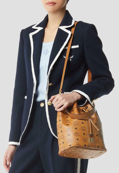 Vrouw in een marineblauwe blazer met witte bies, lichtblauwe top, draagt een beige leren bucketbag met logopatroon over de schouder.