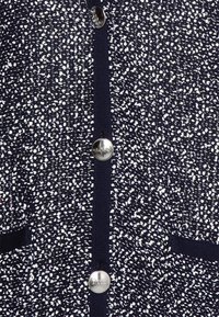 Granatowy kardigan z teksturowanej tkaniny z białymi cętkami, zapinany z przodu na guziki ze srebrnym logo "BOSS" oraz dwoma kieszeniami.