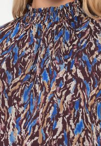 Blusa de cuello alto con un patrón abstracto en colores azul, marrón, crema y burdeos, confeccionada en tela ligera.