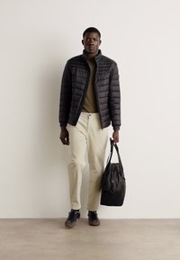 Zwarte pufferjack over olijfgroen coltrui, crèmekleurige broek, bruine sneakers; met een zwarte tas in de hand. Neutrale achtergrond.
