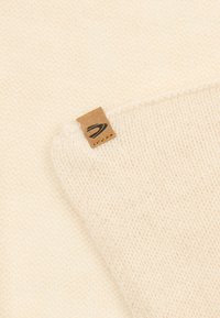 Strickstoff in Cremefarbe mit weicher Textur, an einer Kante mit einem kleinen braunen Leder-Tag versehen, der minimalistisches Design und Materialqualität hervorhebt.