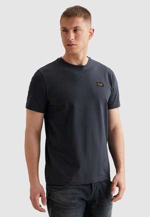 Mand iført en mørkeblå T-shirt med korte ærmer og jeans, der kigger lidt til venstre, står foran en ensfarvet lys baggrund.