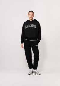 Czarna bluza z kapturem z białym napisem "LAMANIA", kieszeń z przodu oraz czarne dresy z dopasowanym logo. Model nosi srebrne buty sportowe.