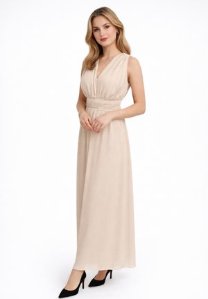Joven mujer con un vestido largo sin mangas de color beige claro y cintura fruncida, llevando tacones negros, de pie con las manos entrelazadas.