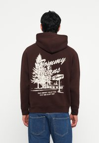 Bruine hoodie met een trekkoordkap, voorzien van een witte grafische afbeelding van bomen en een gebouw op de achterkant. Geribbelde manchetten en onderkant.