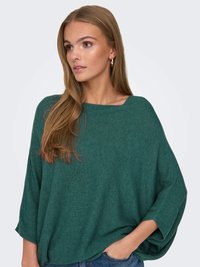 Vihreä ribbitoiminta pullover laajalla kaula-aukolla ja lepakkohihalla. Kangas näyttää pehmeältä ja teksturoidulta. Malli yhdistää sen sinisiin farkkuihin.
