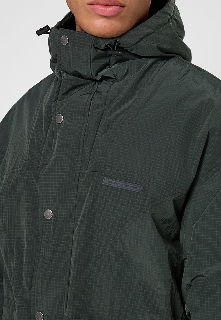 Mężczyzna w ciemnozielonej kurtce z kapturem, zapinanej na zatrzaski, z metką "Element Skateboard Co." na piersi.
