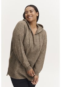 Un hoodie en tricot beige présentant un motif texturé, avec un cordon de serrage et des fentes sur les côtés. Il a une coupe décontractée et des poignets côtelés.