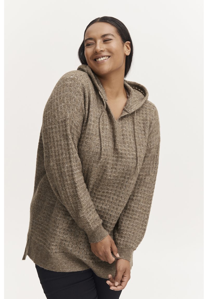 Un hoodie en tricot beige présentant un motif texturé, avec un cordon de serrage et des fentes sur les côtés. Il a une coupe décontractée et des poignets côtelés.