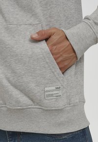 Sudadera gris de tejido suave de mezcla de algodón. Cuenta con un bolsillo frontal, puños acanalados y una etiqueta con instrucciones de cuidado en el costado.