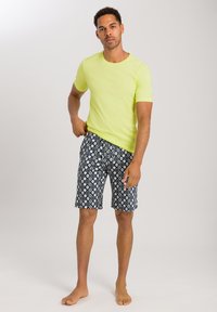 Neon gul kortärmad t-shirt i kombination med mönstrade marinblå shorts med geometriska former. Barfota, med en neutral bakgrund.