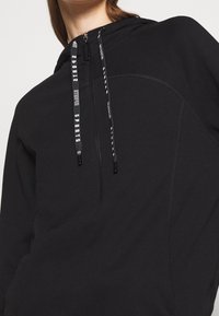 Sudadera negra con capucha y cierre de media cremallera, que presenta cordones texturizados en contraste y un sutil detallado de costuras. Tejido suave.