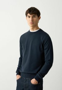 Marineblaues Rundhals-Sweatshirt aus glattem Stoff, mit langen Ärmeln und gerippten Bündchen. Getragen über einem weißen Unterhemd, kombiniert mit dunklen Jeans.