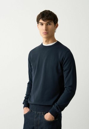 Sweatshirt bleumarin cu guler rotund, realizat dintr-un material neted, cu mâneci lungi și mansete ribbrate. Purtați peste un tricou alb, asortat cu blugi închiși la culoare.