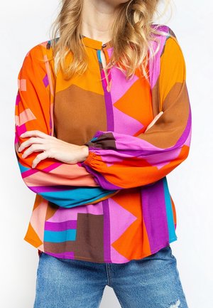 Mujer con los brazos cruzados que lleva una blusa de manga larga geométrica y colorida y vaqueros azules sobre un fondo blanco.