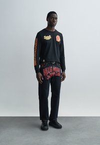 Μαύρο πουκάμισο μακρυμάνικο με κόκκινα γραφικά στα μανίκια, συνδυασμένο με μαύρο τζιν με κόκκινο κείμενο. Φορεμένο με μαύρα αθλητικά παπούτσια.